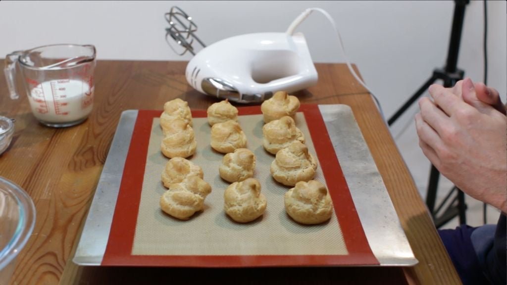 Freshly baked cream puffs resting on a silicone-mat lined cookie sheet on a table. 