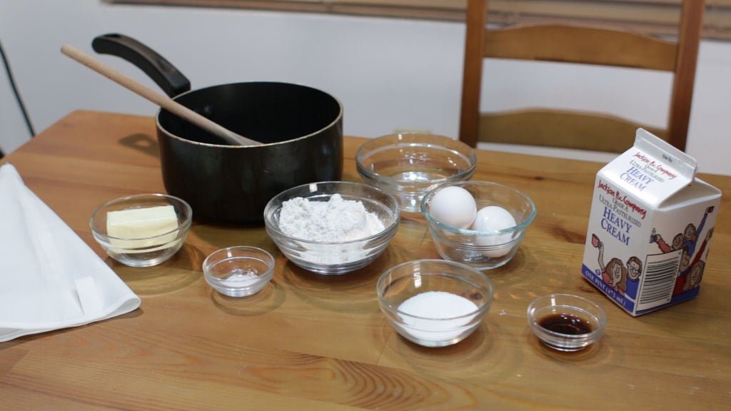 tools and ingredients needed to make the easy cream puffs recipe, eggs, flour, etc. on a wooden table.